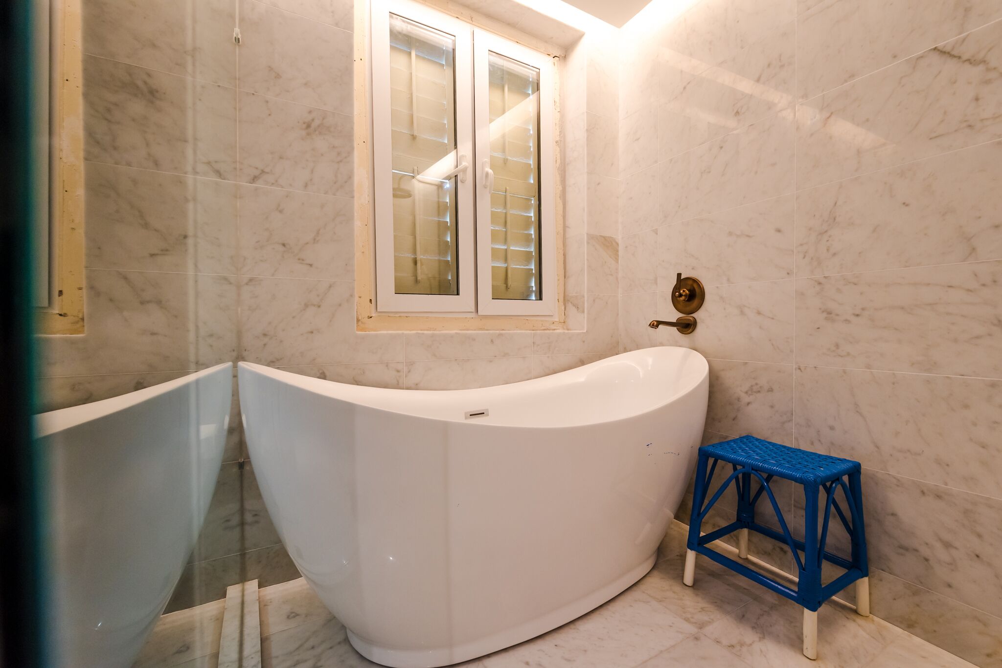 A bathroom with a white bathtub, a window with blinds, and a blue stool.