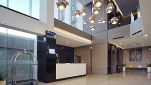 Un hall d’hôtel moderne avec un comptoir d’accueil, un mur de verre et un plafond haut orné de luminaires sphériques suspendus.