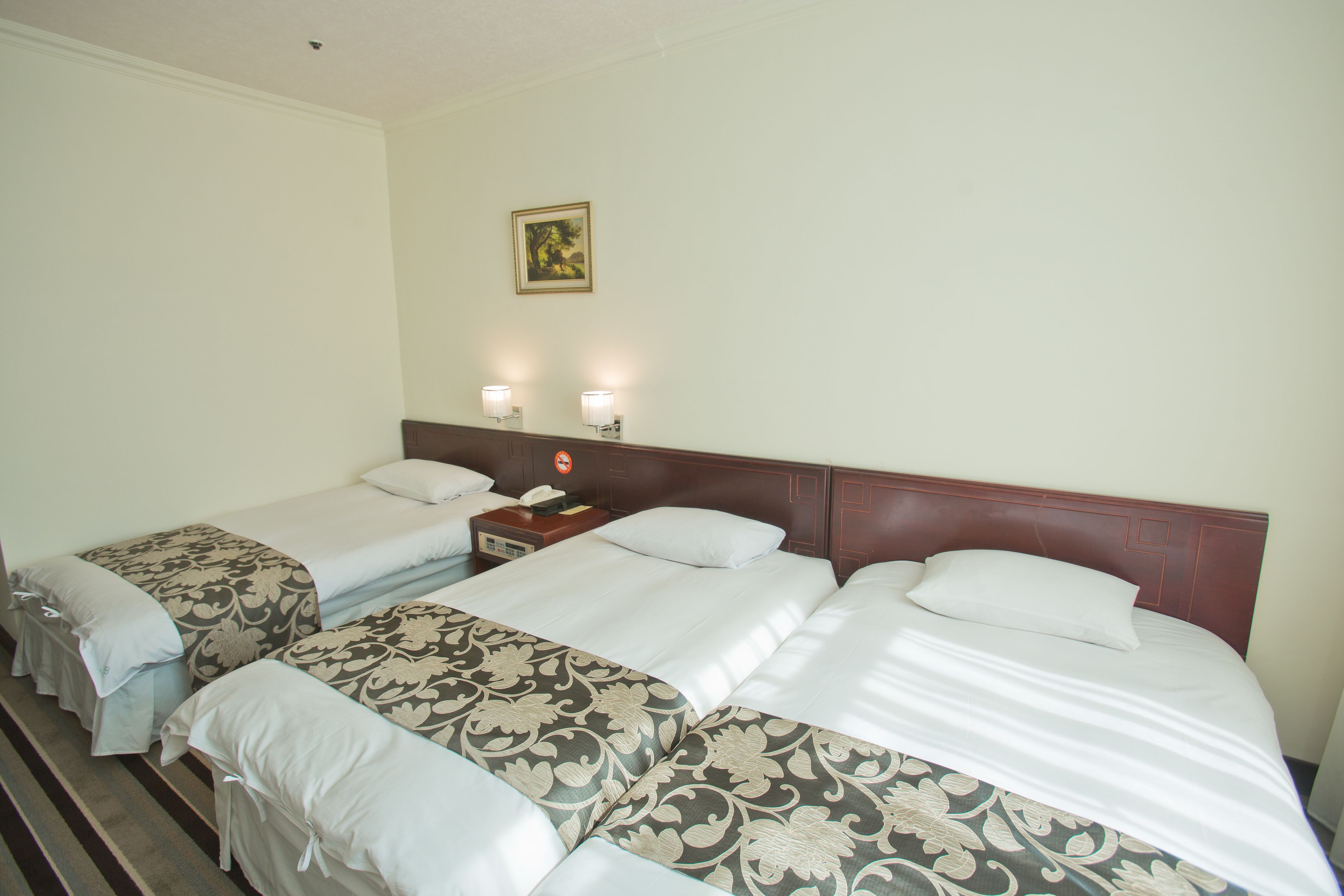 A hotel room with two single beds and one double bed, each with white linens and floral-patterned bedspreads. There are two wall-mounted nightstands with lamps, a telephone, and a small table. A framed picture hangs on the wall above the double bed.