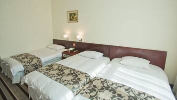 Habitación de hotel con dos camas individuales y una cama doble, cada una con sábanas blancas y colchas estampadas con motivos florales. Hay dos mesitas de noche fijadas a la pared con lámparas, un teléfono y una pequeña mesa. Encima de la cama doble cuelga un cuadro enmarcado.