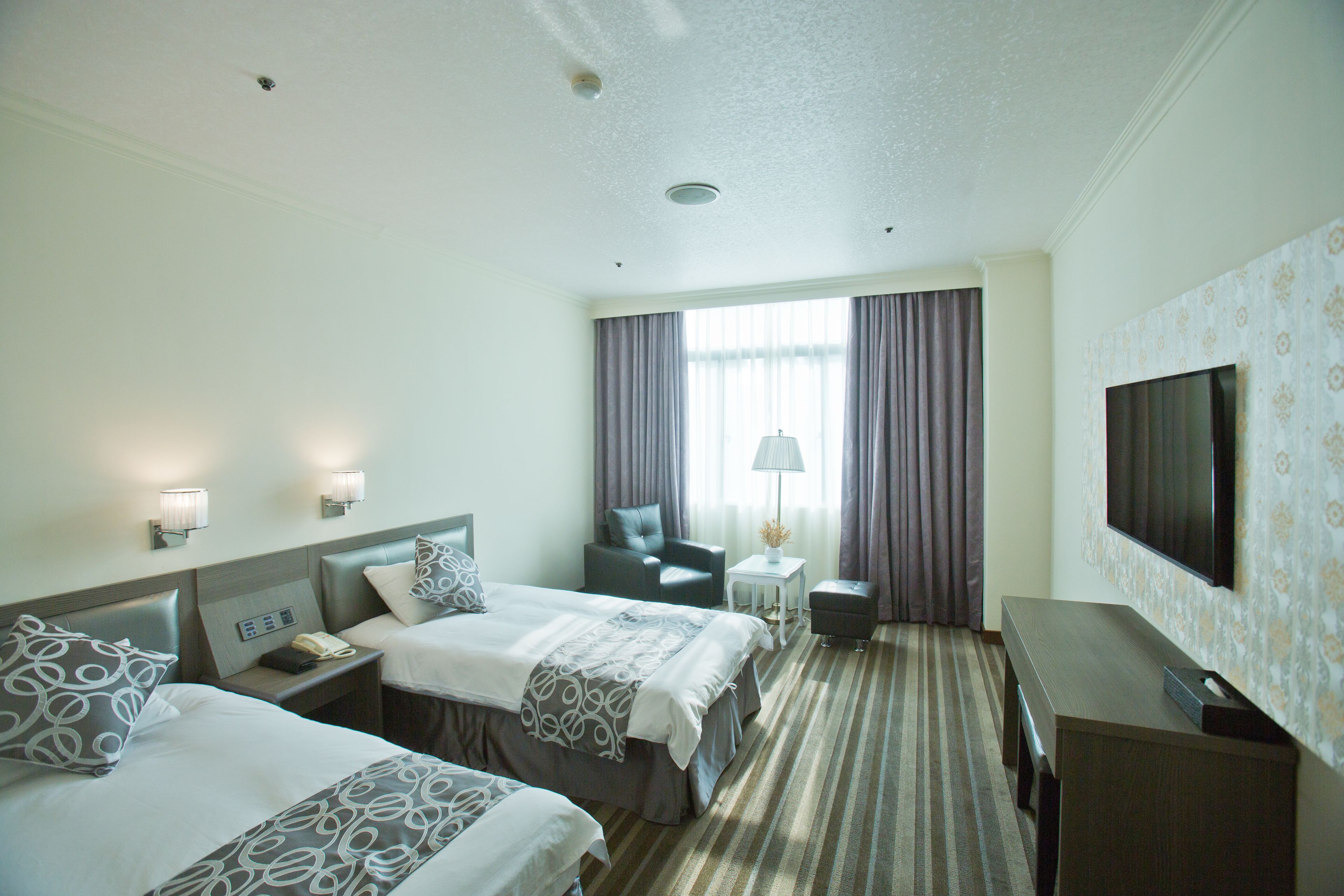 A hotel room with two beds, a desk, a chair, a TV, and a lamp.