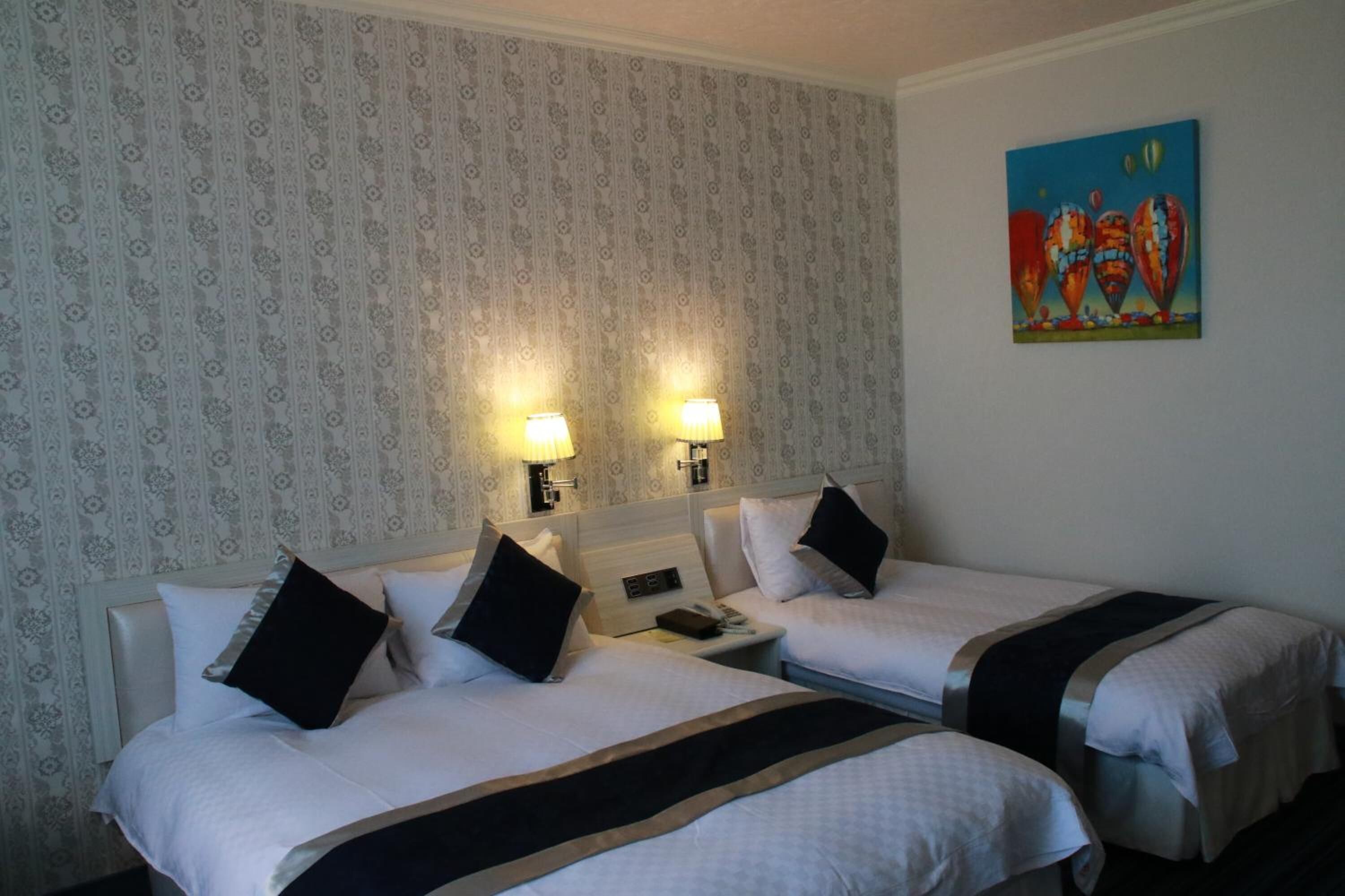 A hotel room with two beds, each with a headboard, bedside tables, lamps, and a painting on the wall.