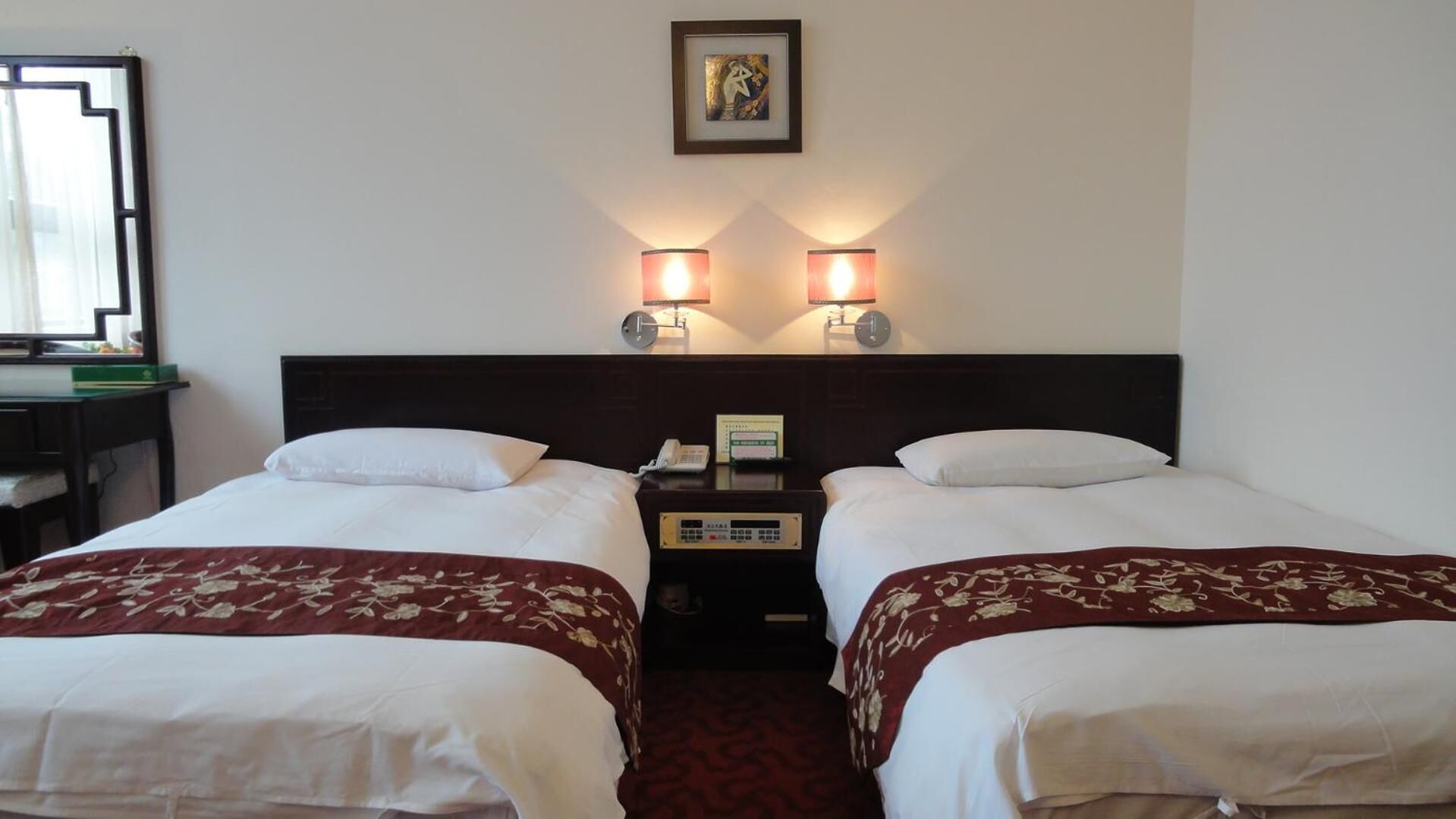 A hotel room with two beds, each with white linens and a red floral bedspread. There are two wall-mounted lamps, a framed picture above the beds, and a small table with a telephone and a clock.
