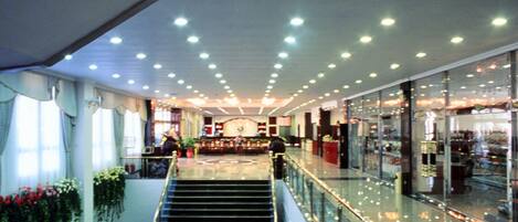 A multi-level shopping mall with a central staircase, glass railings, and various shops on the upper level.