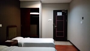 Ein Hotelzimmer mit zwei Betten, einem hölzernen Nachttisch, einer Tür mit rotem Rahmen und einem Lichtschalter an der Wand.