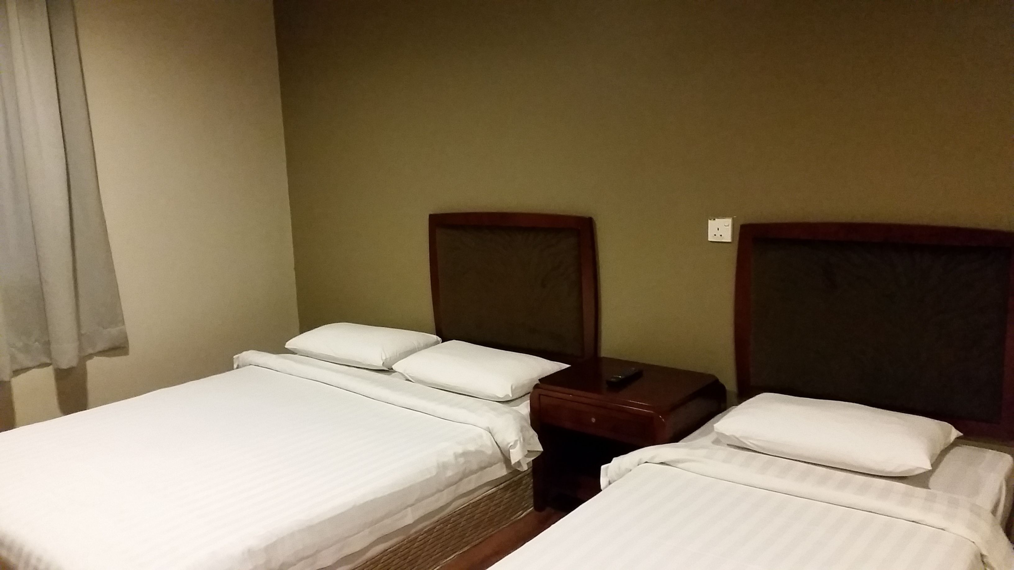 A hotel room with two beds, each with white linens and pillows, a wooden headboard, a nightstand with a lamp, and a power outlet on the wall.