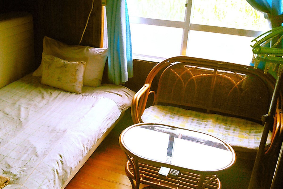 Une petite pièce agréable, dotée d’un lit simple, d’une chaise avec une table et d’une fenêtre avec des rideaux.