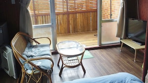 Ein Zimmer mit Bett, Sessel, kleinem Tisch, Fernseher und einer Glastür zum Außenbereich mit Holzterrasse.