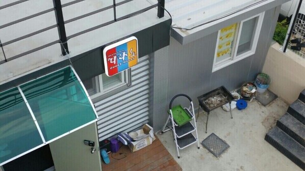 A small, single-story building with a sign indicating it is a guest house. There is a small outdoor area with a metal chair and a trash bin.