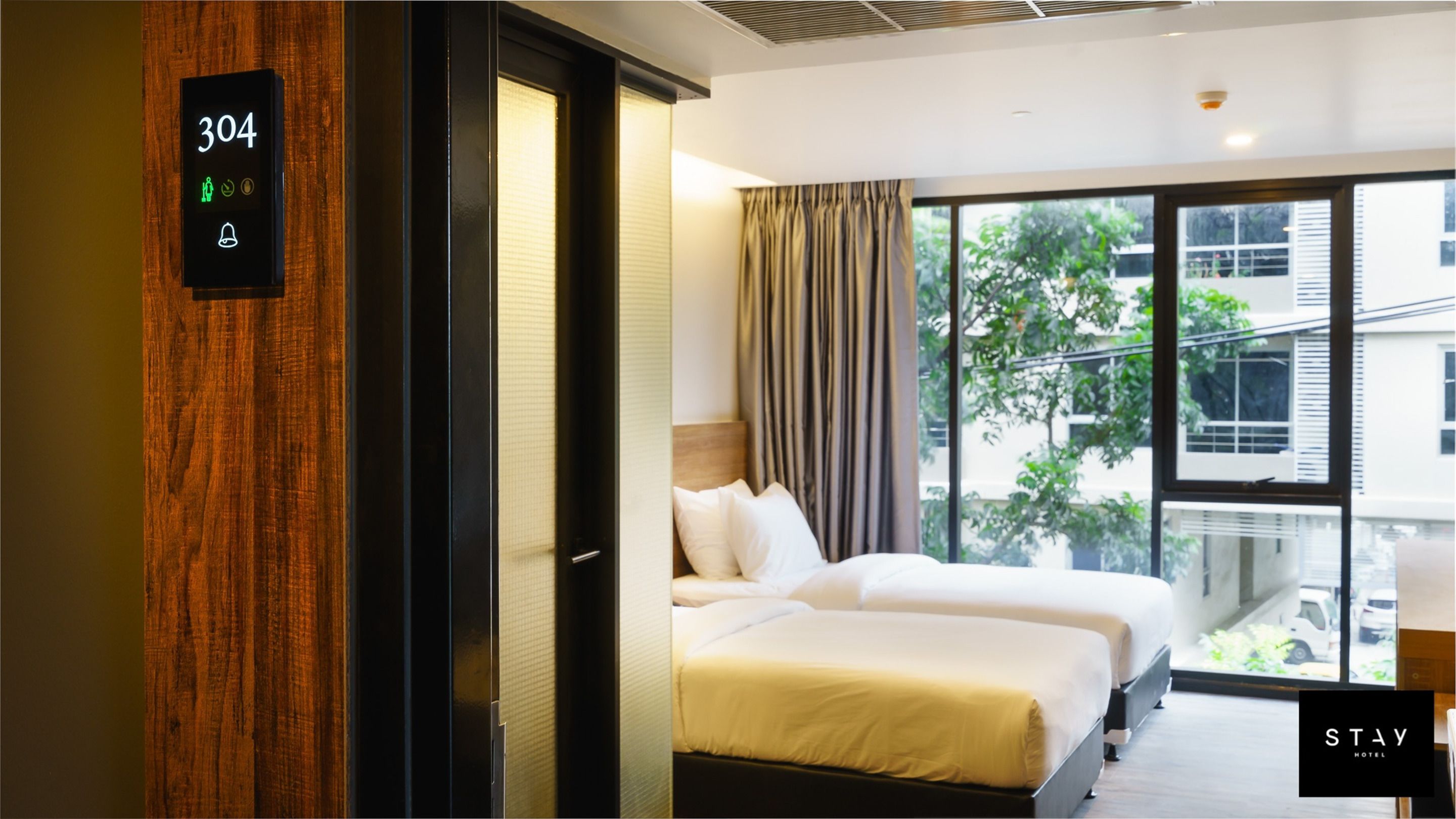 A hotel room with two beds, a digital display showing room number 304, and a view of the outside through large windows.