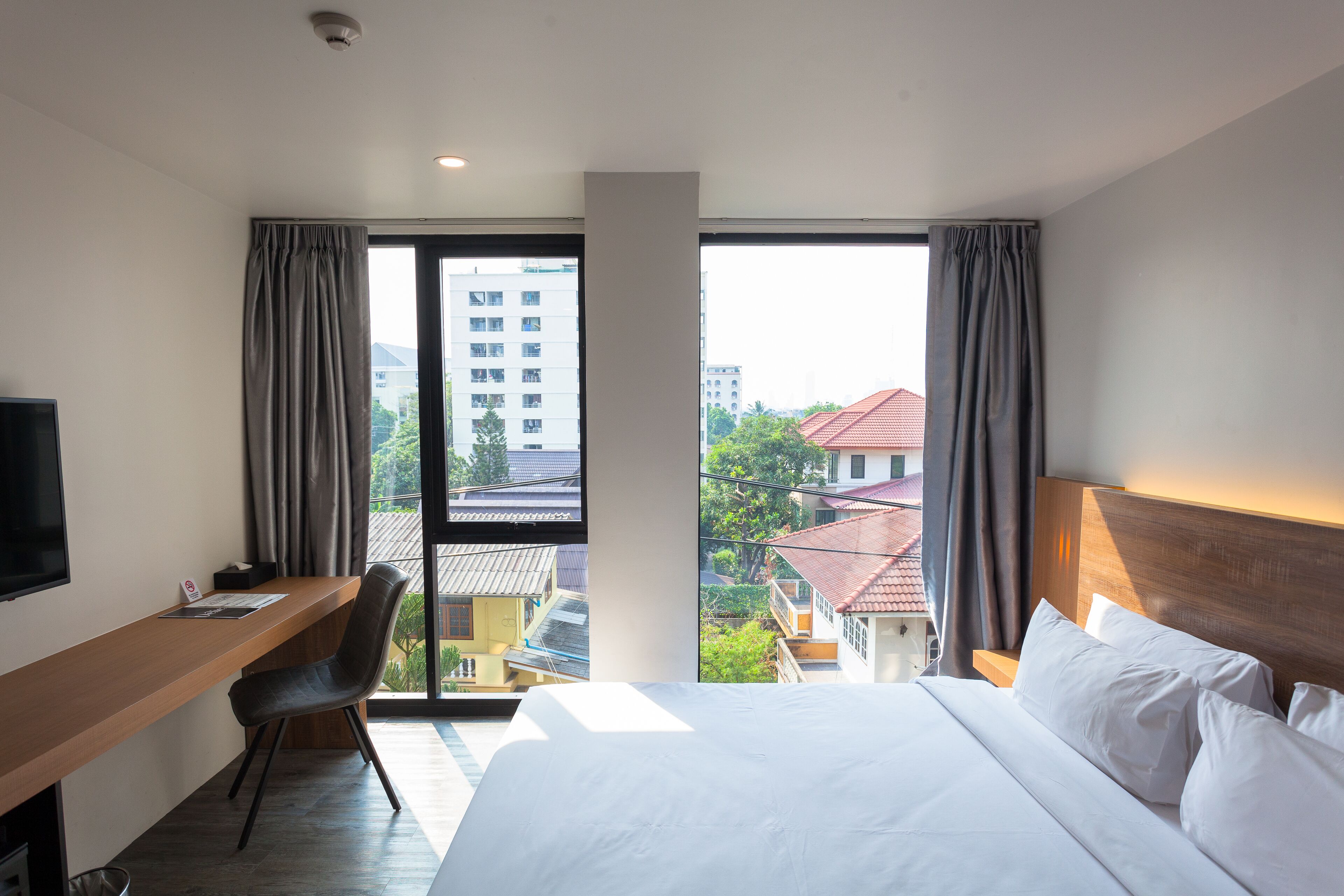A hotel room with a bed, a desk, a chair, a television, and large windows offering a view of buildings and trees.