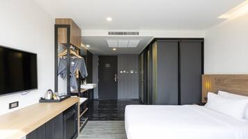 A modern hotel room with a flat-screen TV, a wooden desk with a sink, a robe hanging on a rack, a bed with white linens, and a wardrobe with sliding doors.