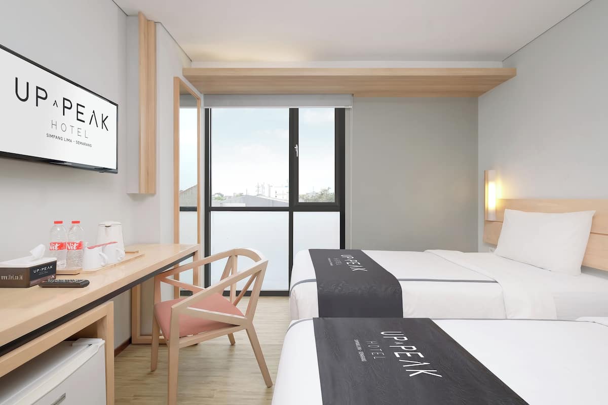 A hotel room with two beds, a desk, and a chair. There is a sign on the wall and a window with a view.