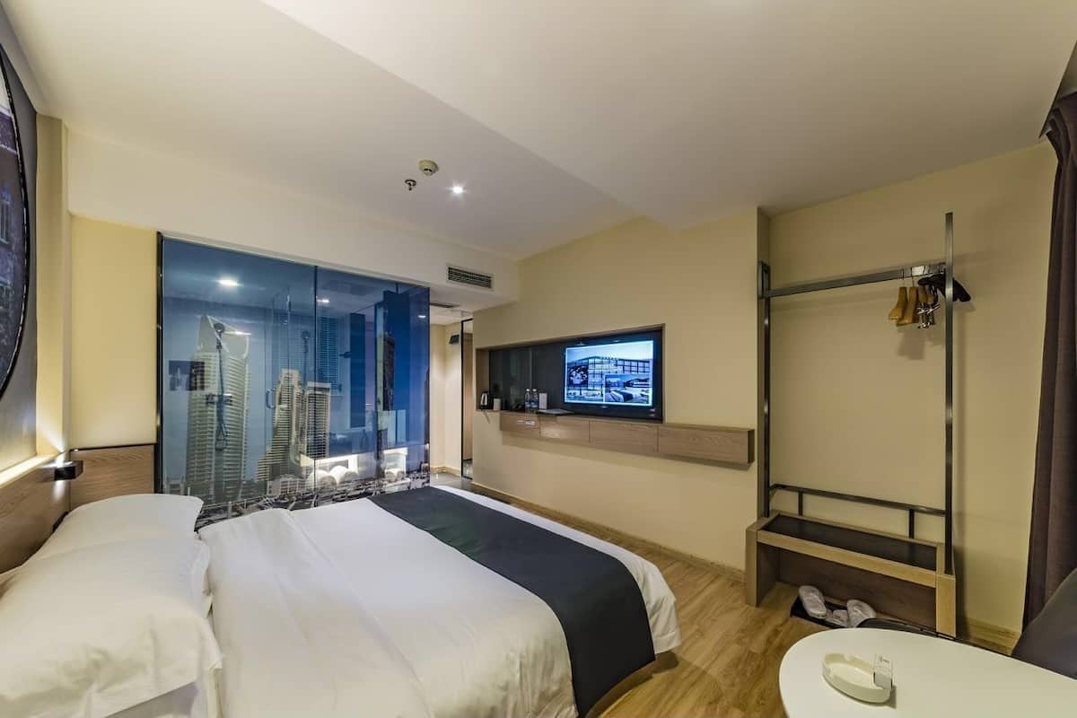 A hotel room with a large bed, a TV mounted on the wall, a shoe rack, and a view of the city through a glass door.