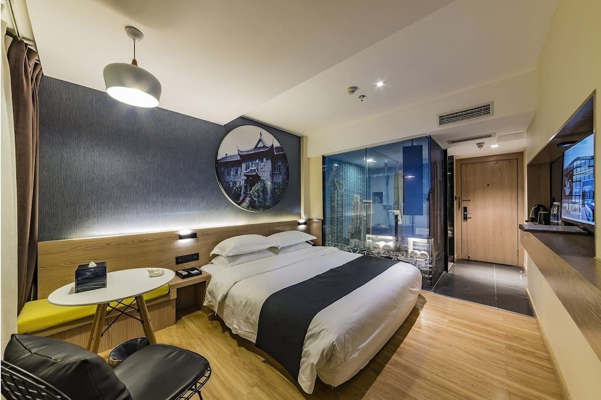 A modern hotel room with a bed, bedside table, chair, and a large window offering a city view.