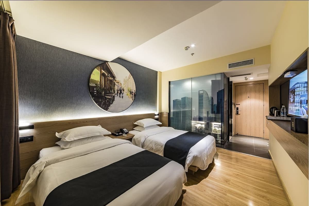 A hotel room with two beds, a large mirror, a cityscape view, a wooden door, and a small desk with a TV.
