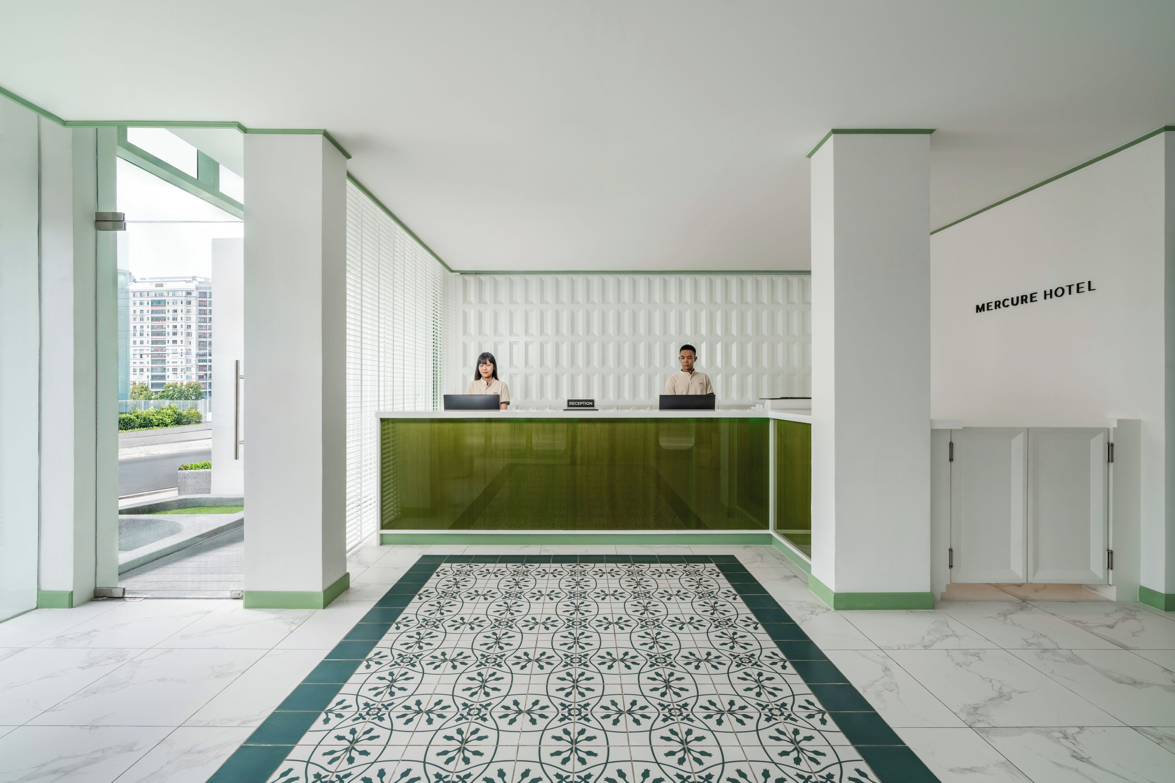 Un espace d’accueil d’hôtel comprenant un comptoir de réception, deux réceptionnistes, un sol à motifs et une vue sur l’extérieur.