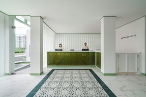 Een hotel receptie met een receptiebalie, twee receptionisten, een vloer met patroon en uitzicht op buiten.