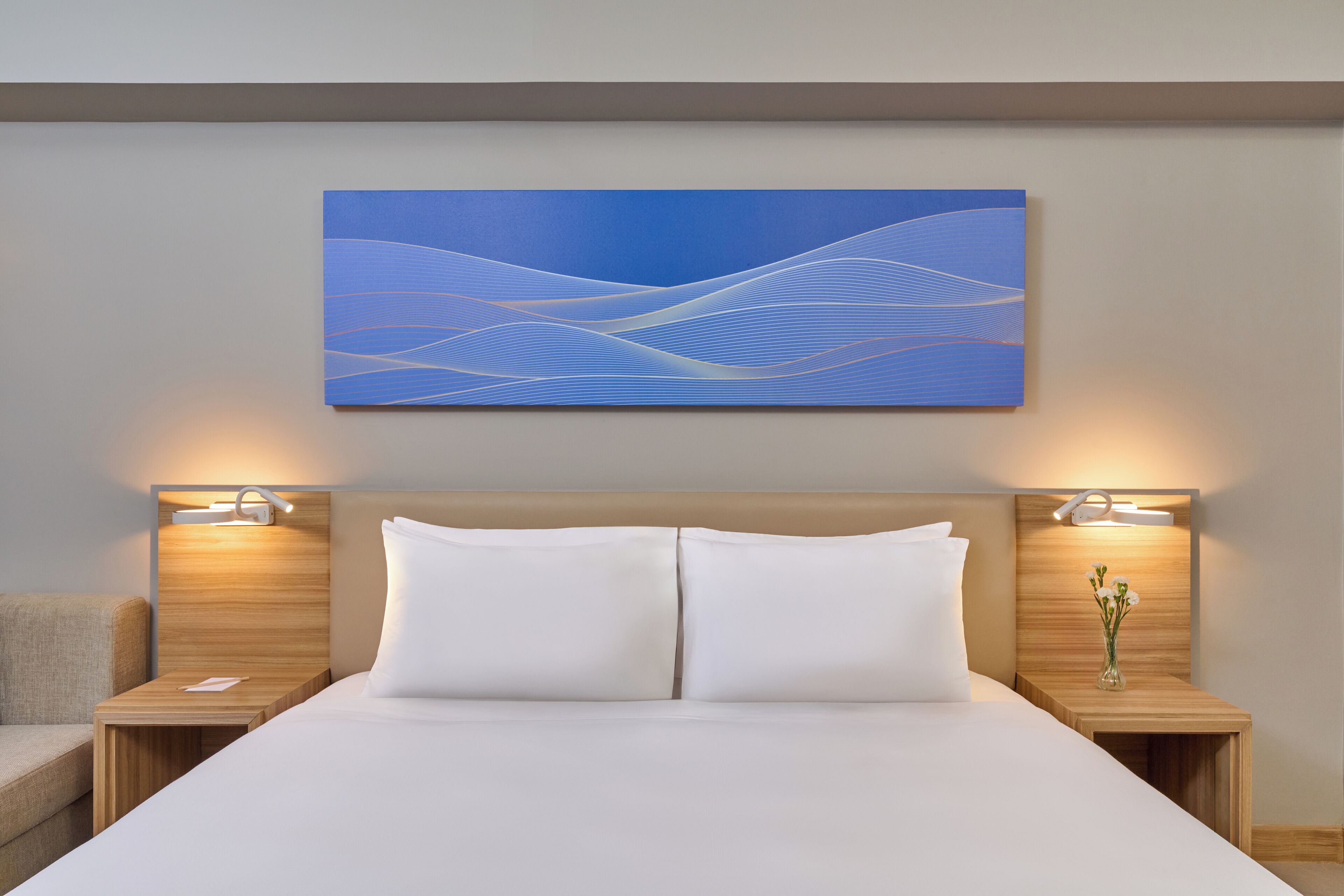 Een moderne hotelkamer met een groot bed, twee nachtkastjes met lampen, een kunstwerk aan de muur en een vaas met bloemen op een van de nachtkastjes.