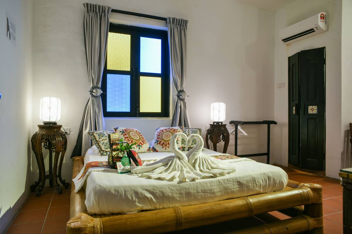 A neatly made bed with a swan towel design, a window with curtains, two bedside tables with lamps, and a door with a decorative panel.