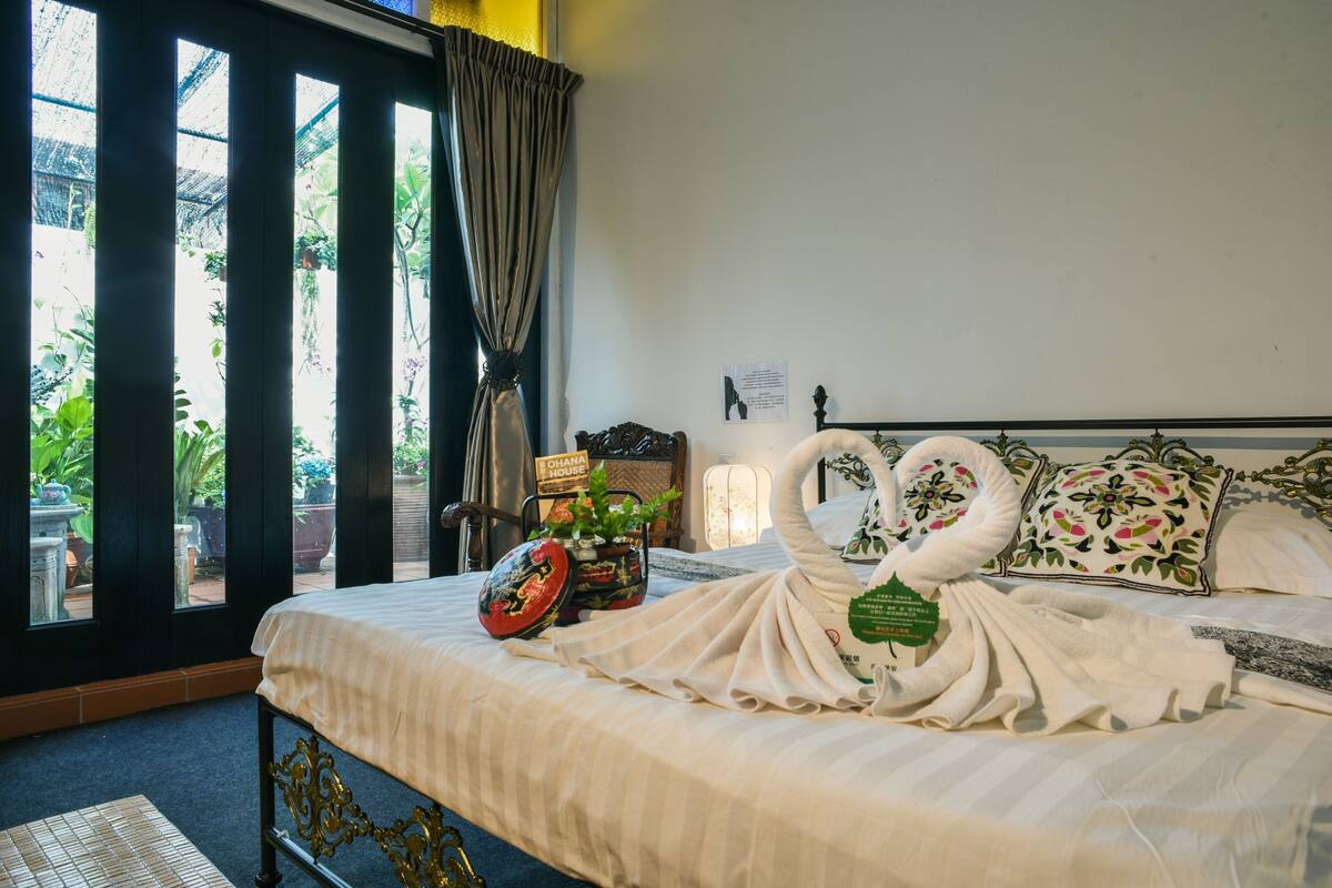 A hotel room with a bed, a potted plant, a sign reading 'OHANA HOUSE', and a view of the outdoors.