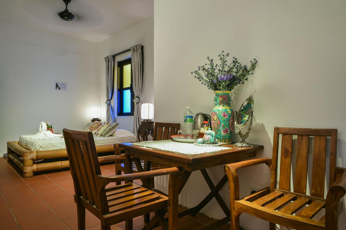 A dining area with a wooden table, chairs, a vase with flowers, and a bed in the background.