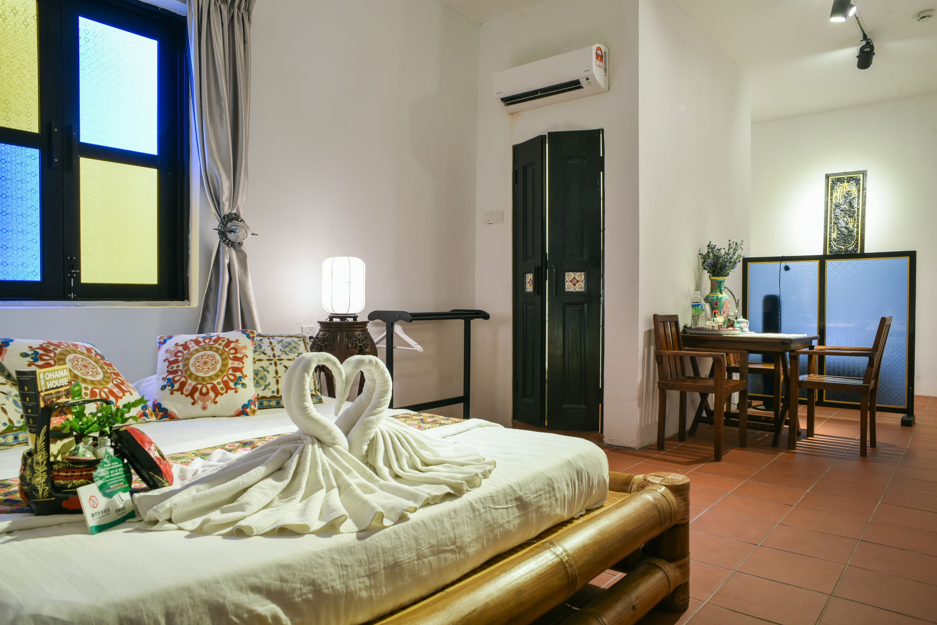 A hotel room with a bed, a dining table, chairs, a decorative swan towel, a window with curtains, an air conditioning unit, and a wall-mounted shelf with a vase.