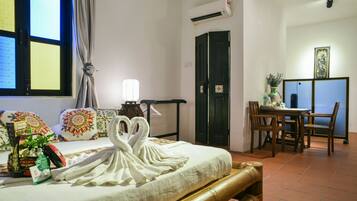 A hotel room with a bed, a dining table, chairs, a decorative swan towel, a window with curtains, an air conditioning unit, and a wall-mounted shelf with a vase.