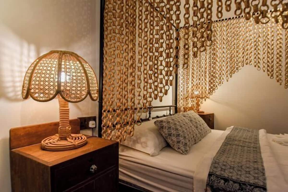 A bed with a patterned blanket, a wooden bedside table with a lamp, and a wall with a hanging chain decoration.
