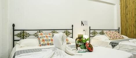 Una habitación de hotel con dos camas, cada una con ropa de cama blanca y almohadas decorativas. Hay un cartel en la pared y una planta al lado izquierdo de la habitación.