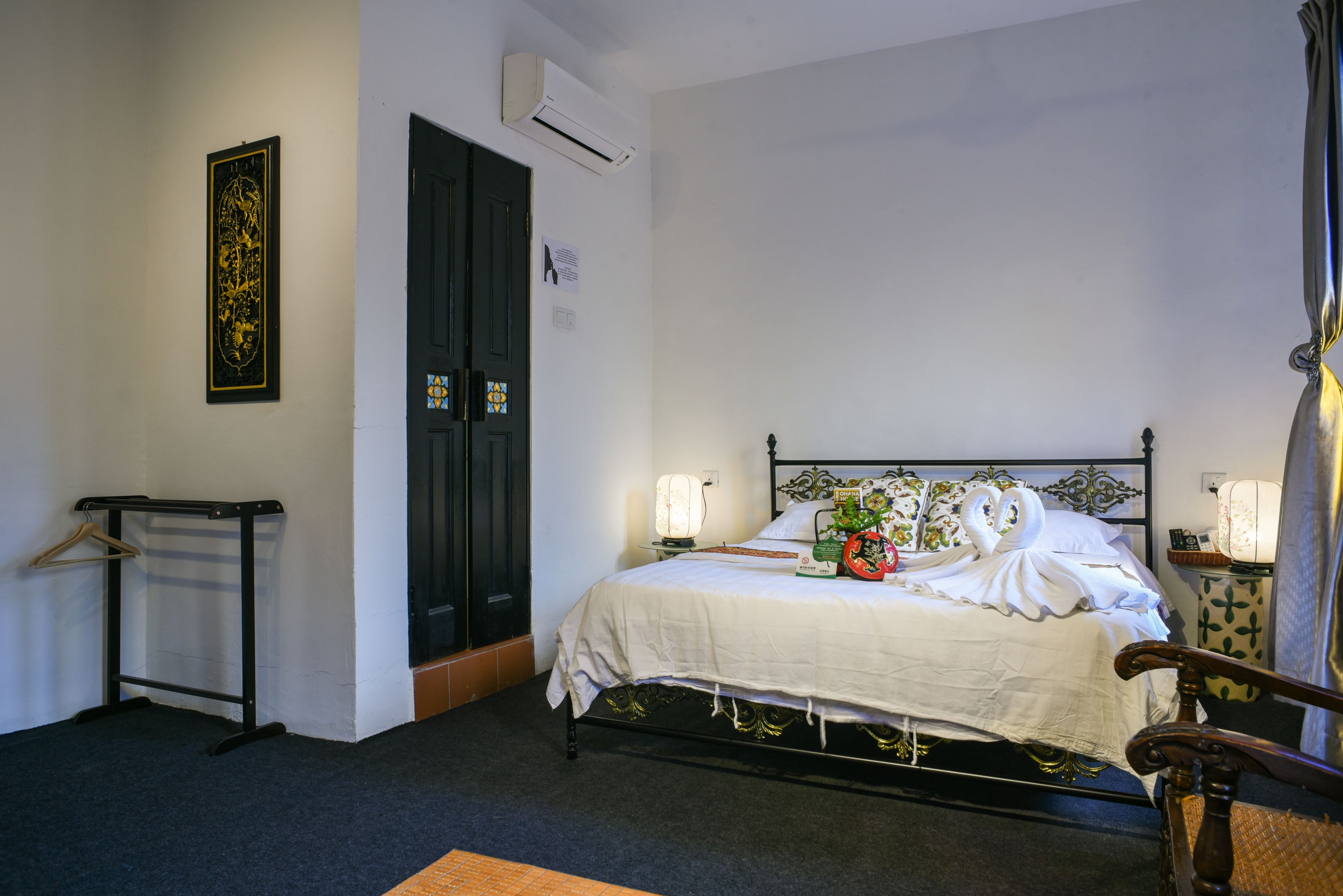Een opgemaakt bed met een witte dekbedovertrek en decoratieve kussens, een nachtkastje met een lamp en een wandairconditioner.