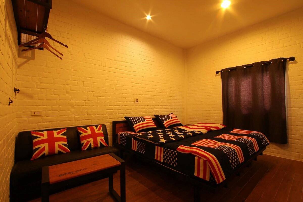 A room with a white brick wall, a wooden bench, and two beds with American flag-themed bedding.