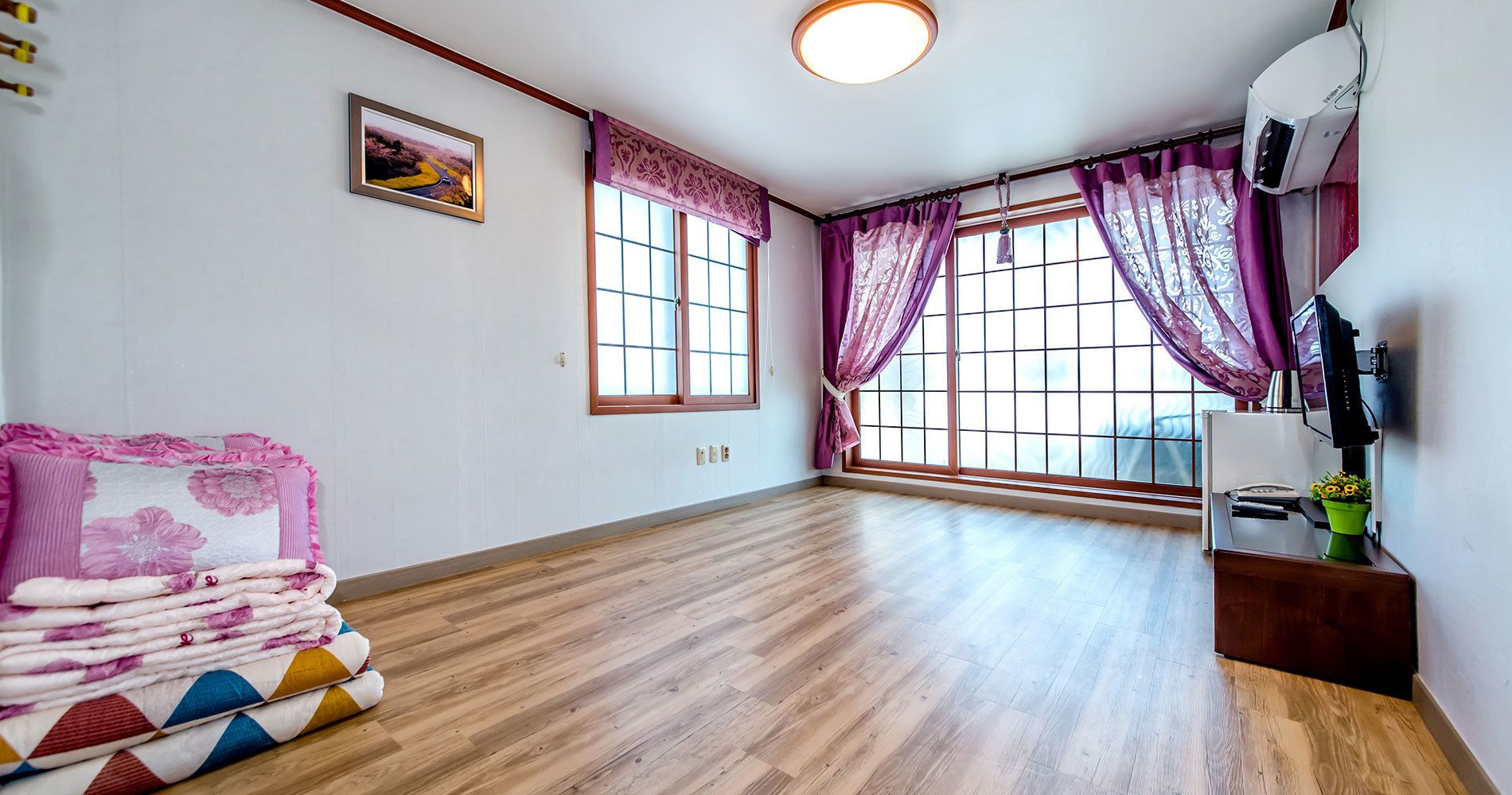 A room with wooden flooring, large windows with purple curtains, a framed picture on the wall, and a small table with a plant.