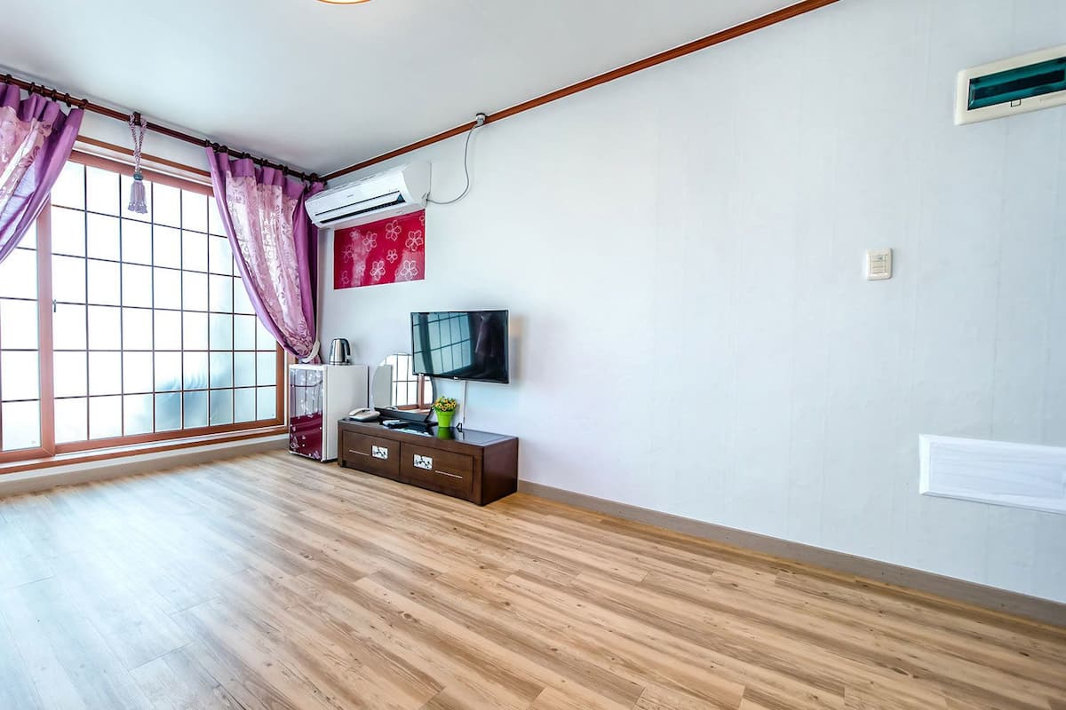 A room with a large window, wooden flooring, a TV, a small cabinet, and an air conditioning unit.