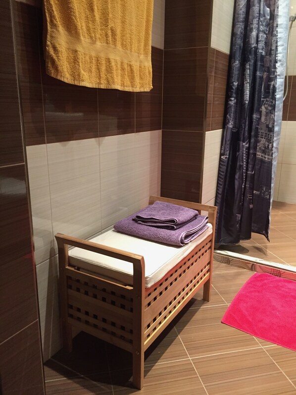 Una cama de madera pequeña con un colchón blanco y una toalla morada encima. Una silla de madera está colocada frente a la cama.