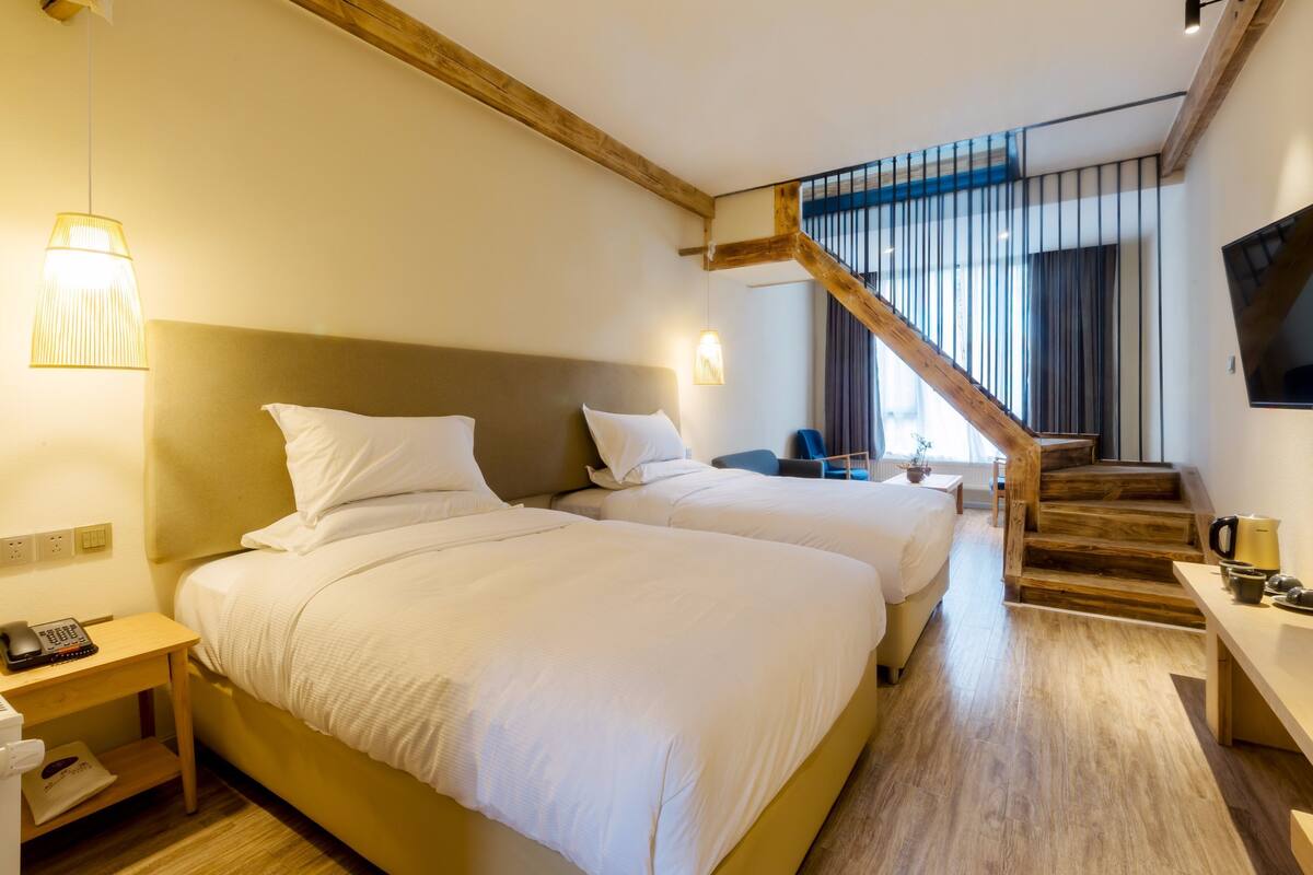 Habitación de hotel con dos camas, una escalera de madera, un escritorio con una tetera y un televisor en la pared.