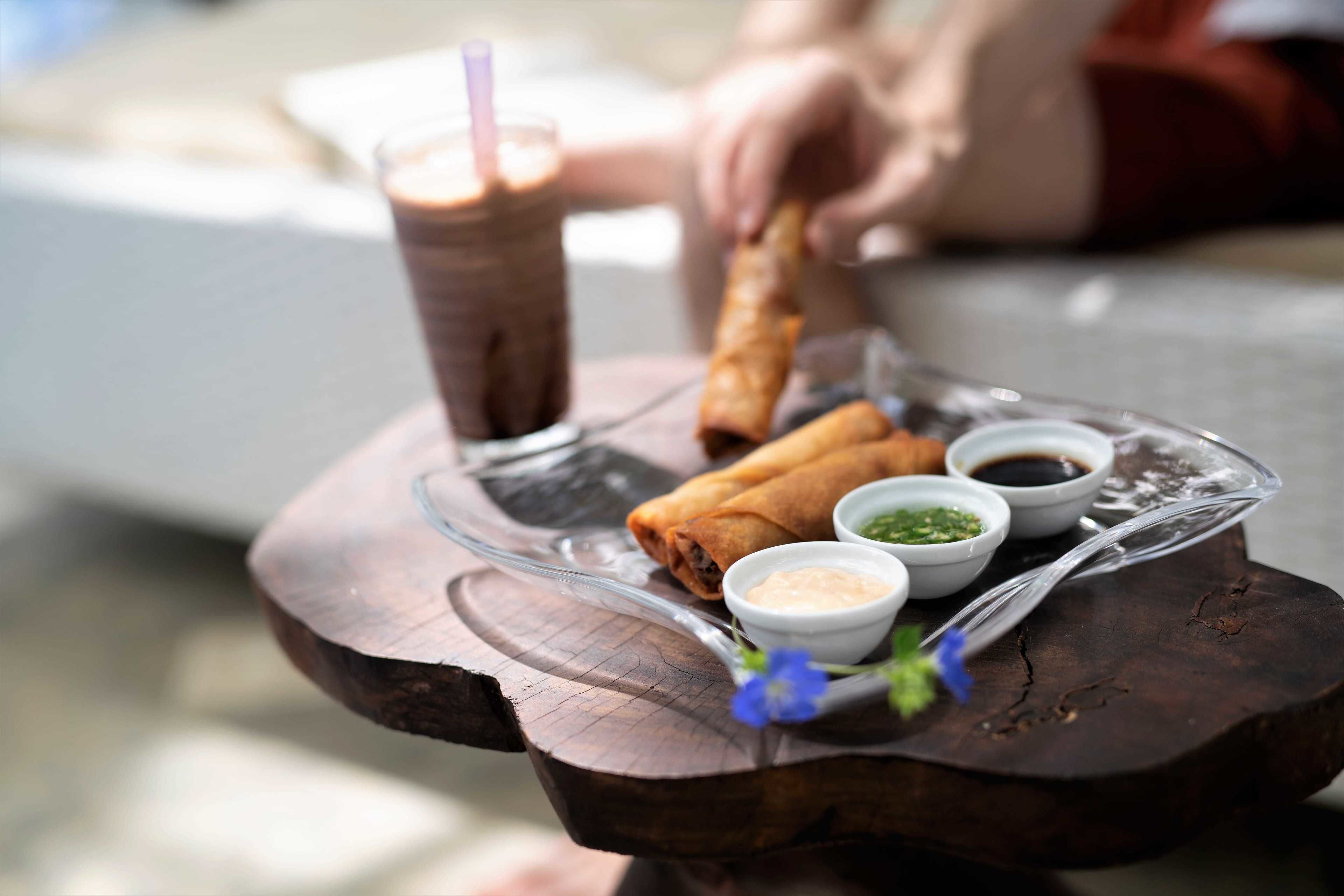 Uma mesa de madeira com um suporte de vidro que contém uma bebida, dois rolinhos primavera e três pequenos recipientes de molhos.