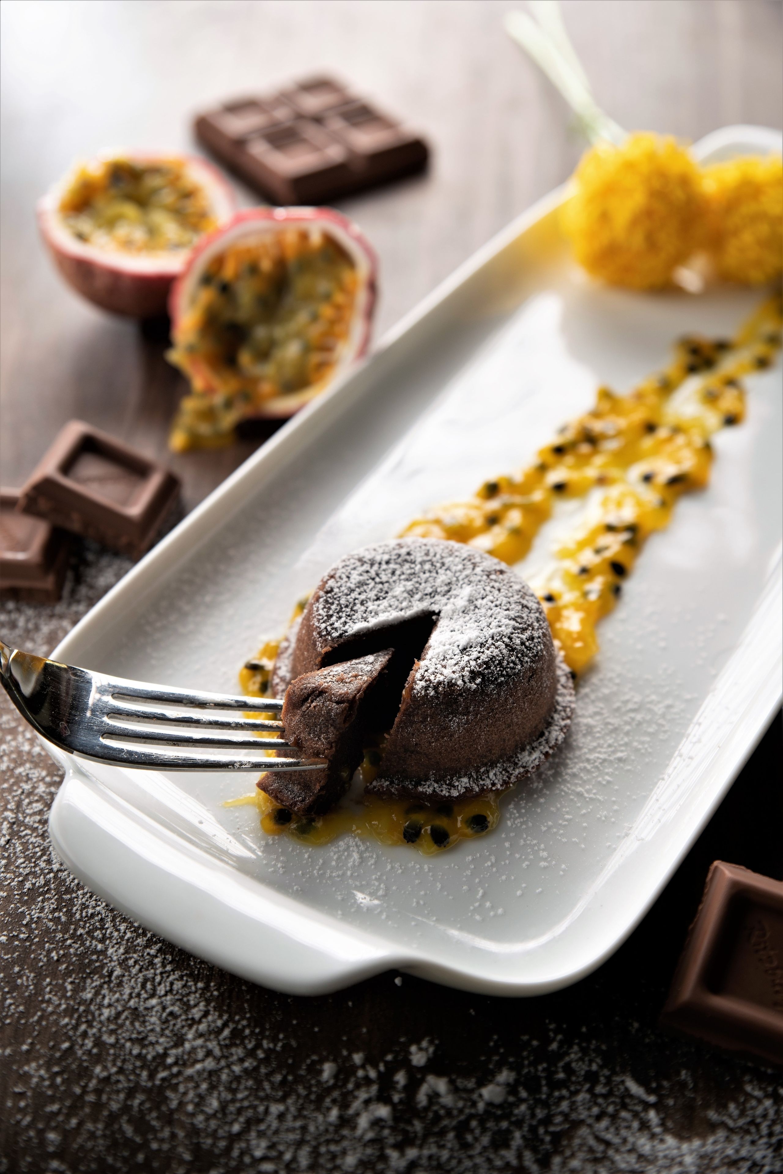 A dessert on a white plate with a fork, surrounded by chocolate pieces and passion fruit halves.