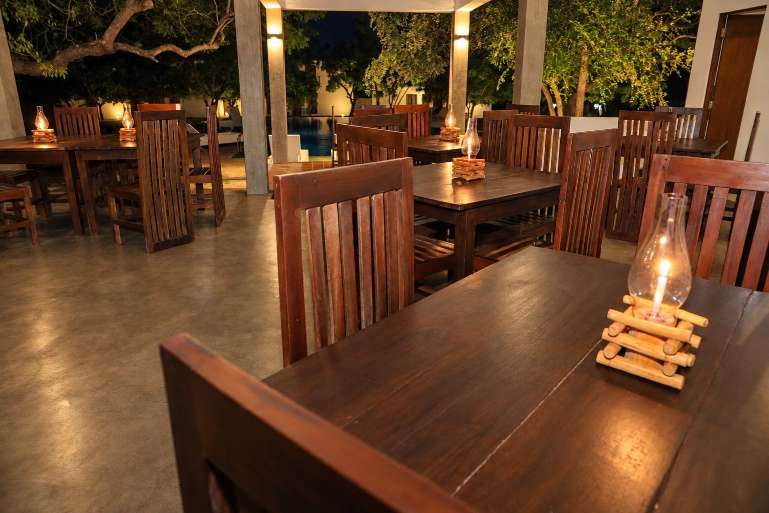 An outdoor dining area with wooden tables and chairs, surrounded by trees and a pool.