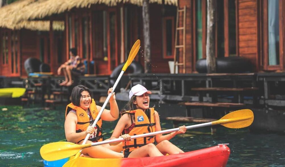 Two people kayaking in a river with thatched-roof buildings in the background.