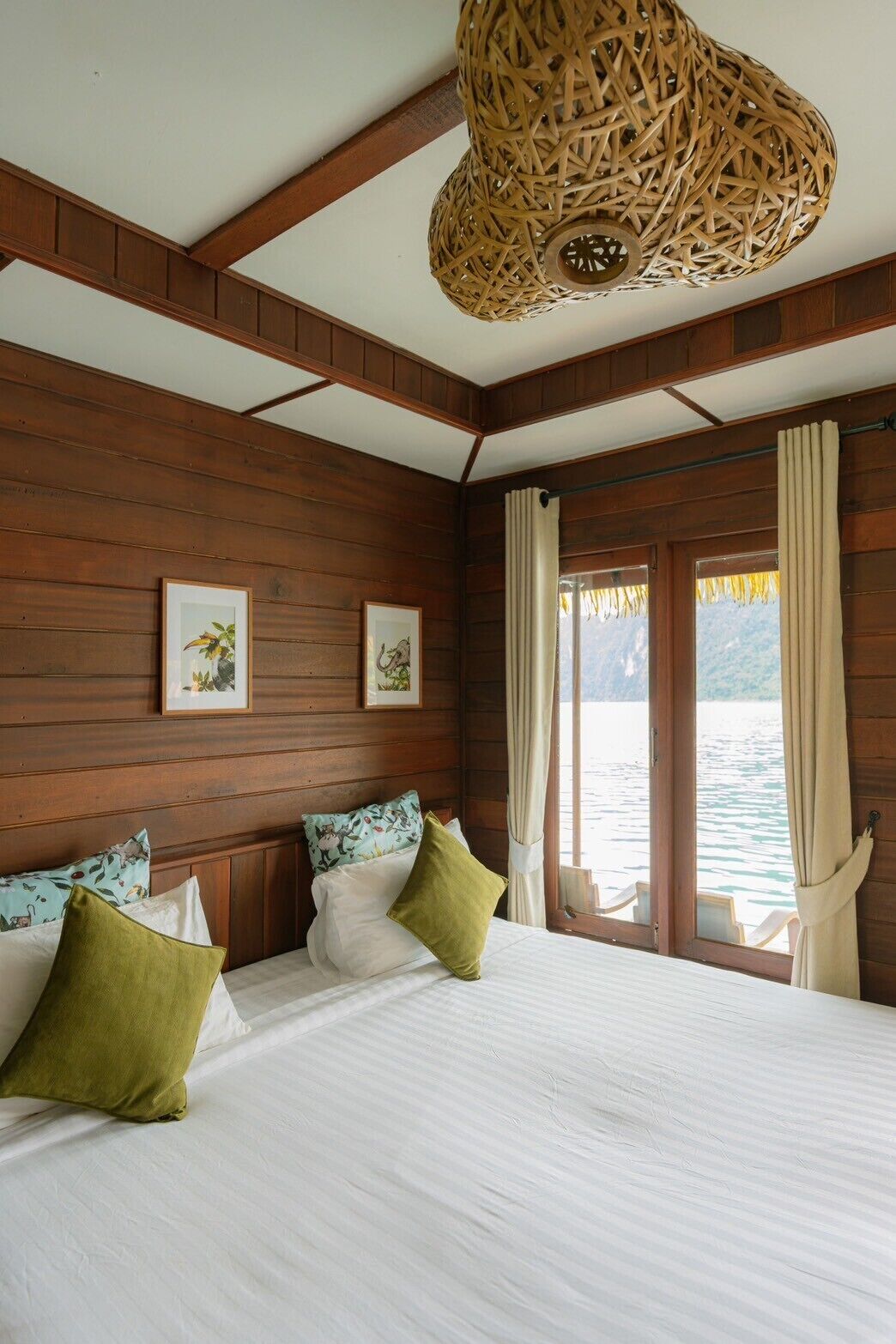 Una habitación tipo cabaña de madera con una cama, dos cuadros enmarcados, una lámpara de techo tejida y una ventana con cortinas.