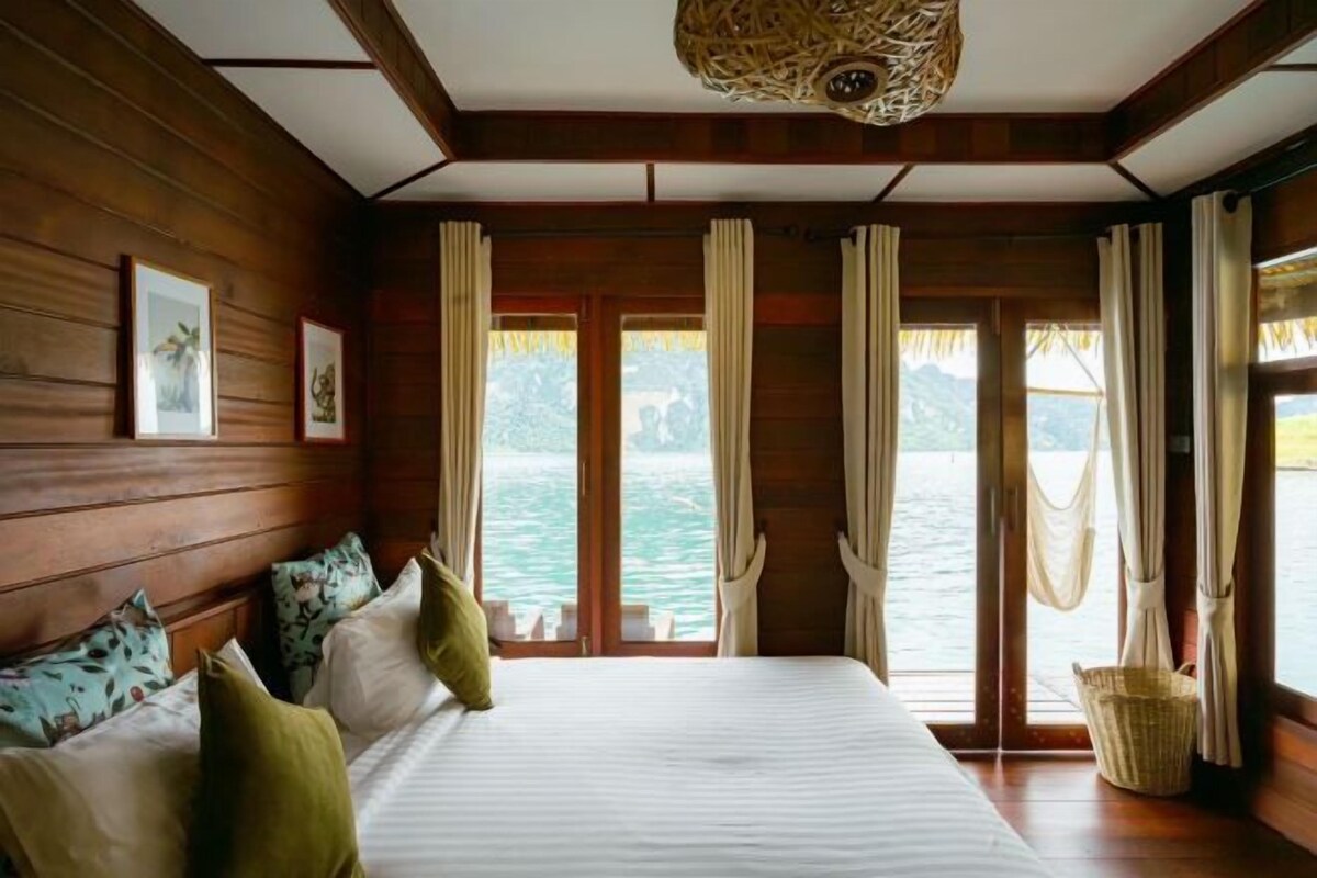 A wooden cabin room with a bed, curtains, and a view of the water.