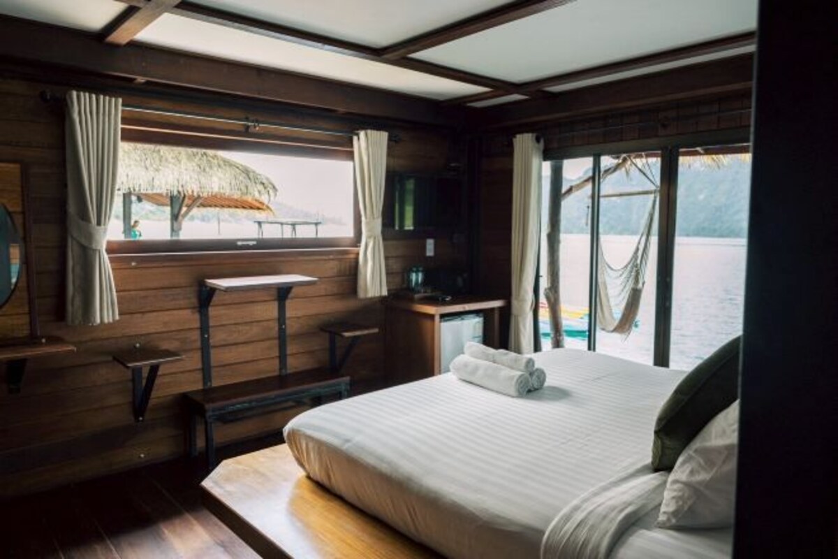 A cabin with a bed, a small table, a bench, a window with curtains, and a view of the water.