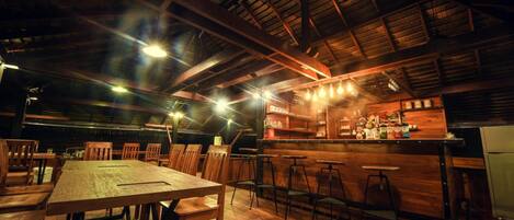 A rustic interior with wooden tables and chairs, a bar area with stools, and a high ceiling with exposed beams.
