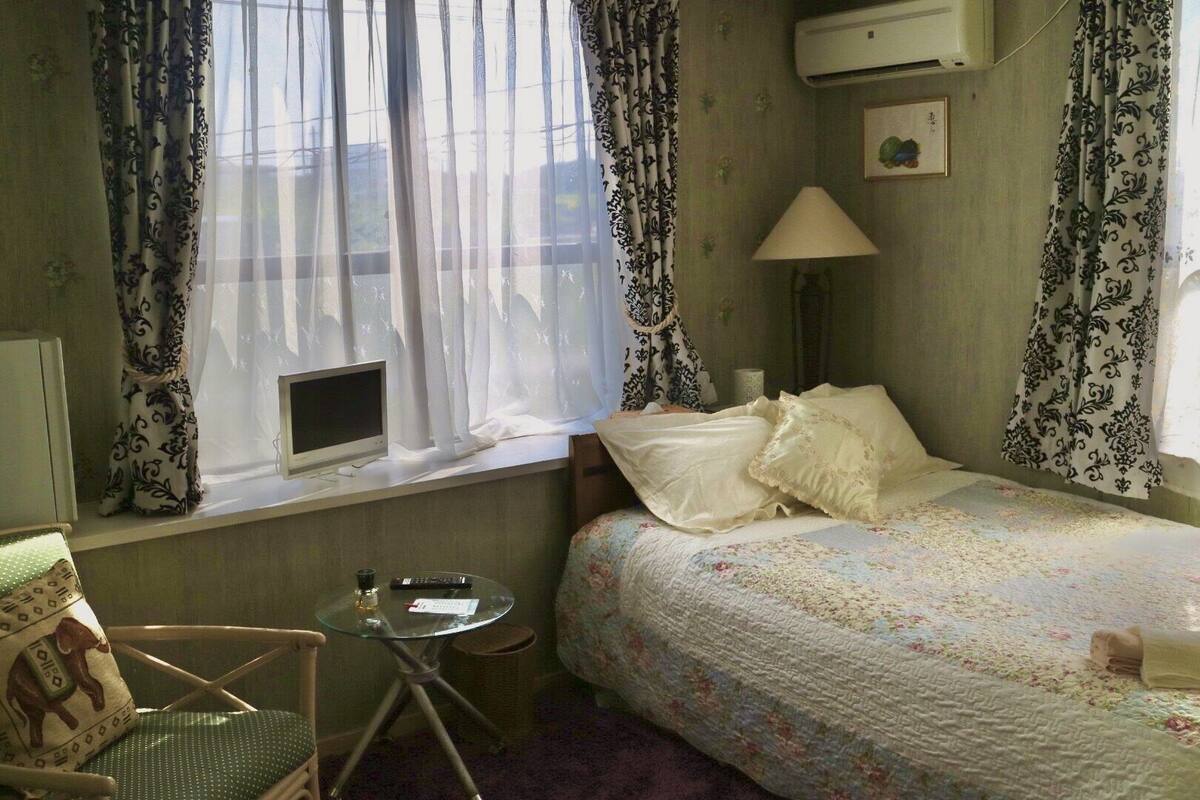 A small, cozy room with a bed, a chair, a small table, and a window with curtains.