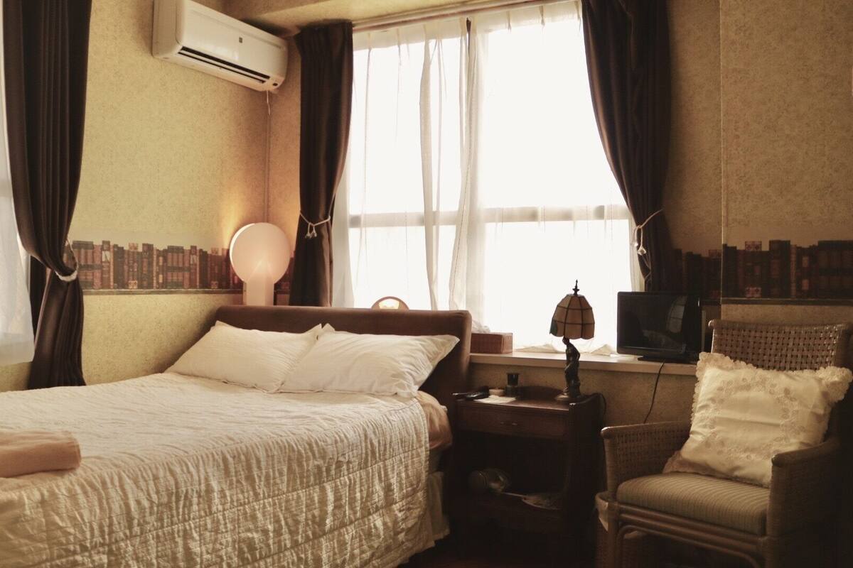 A neatly made bed with white linens, a bedside lamp, a chair with a cushion, and a television on a small table.