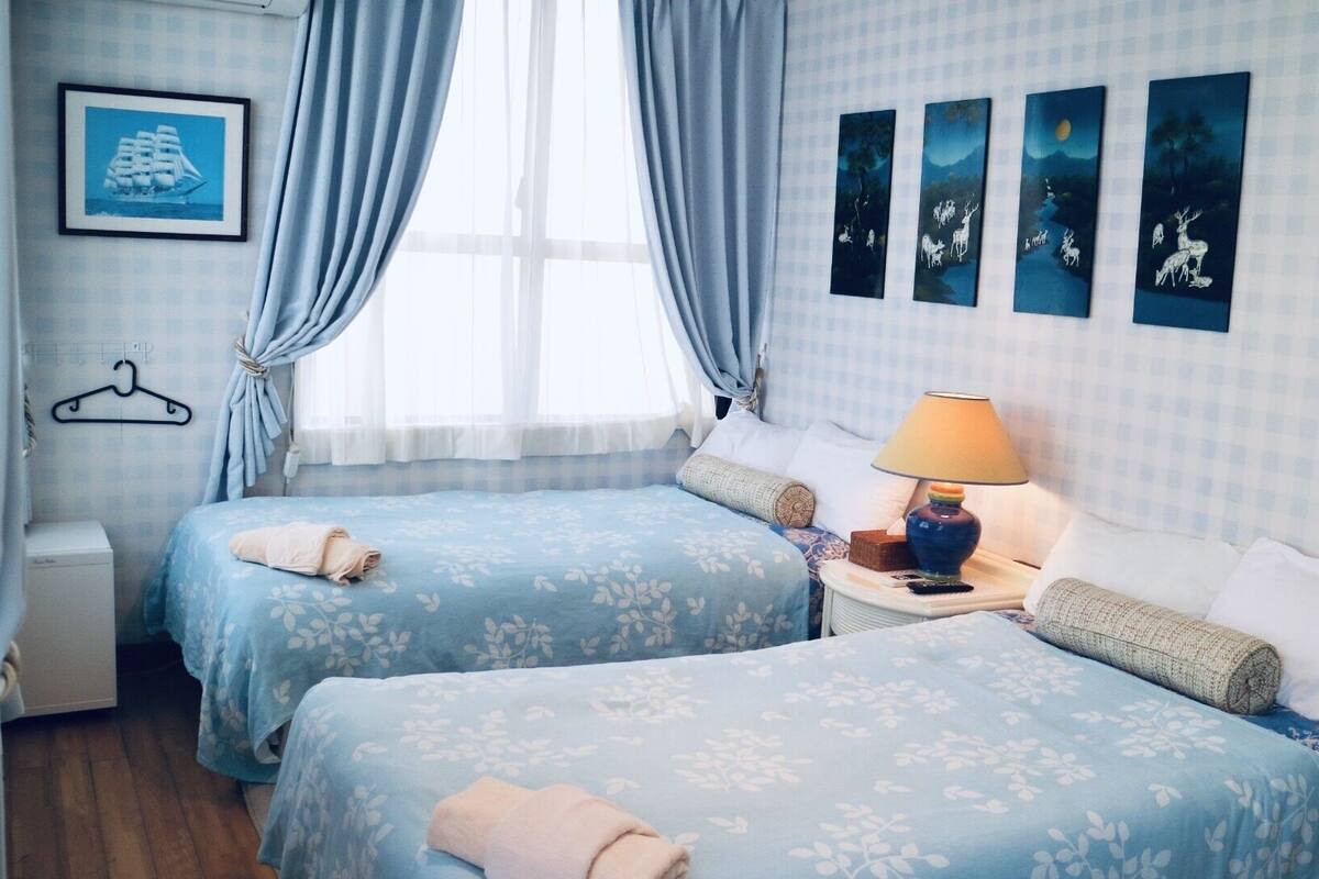 A hotel room with two beds, a nightstand, a lamp, a wall art piece, and a window with curtains.