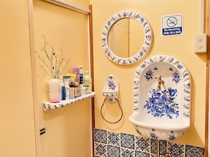 A bathroom with a yellow wall, a no smoking sign, a mirror, a sink with floral designs, and toiletries on a shelf.