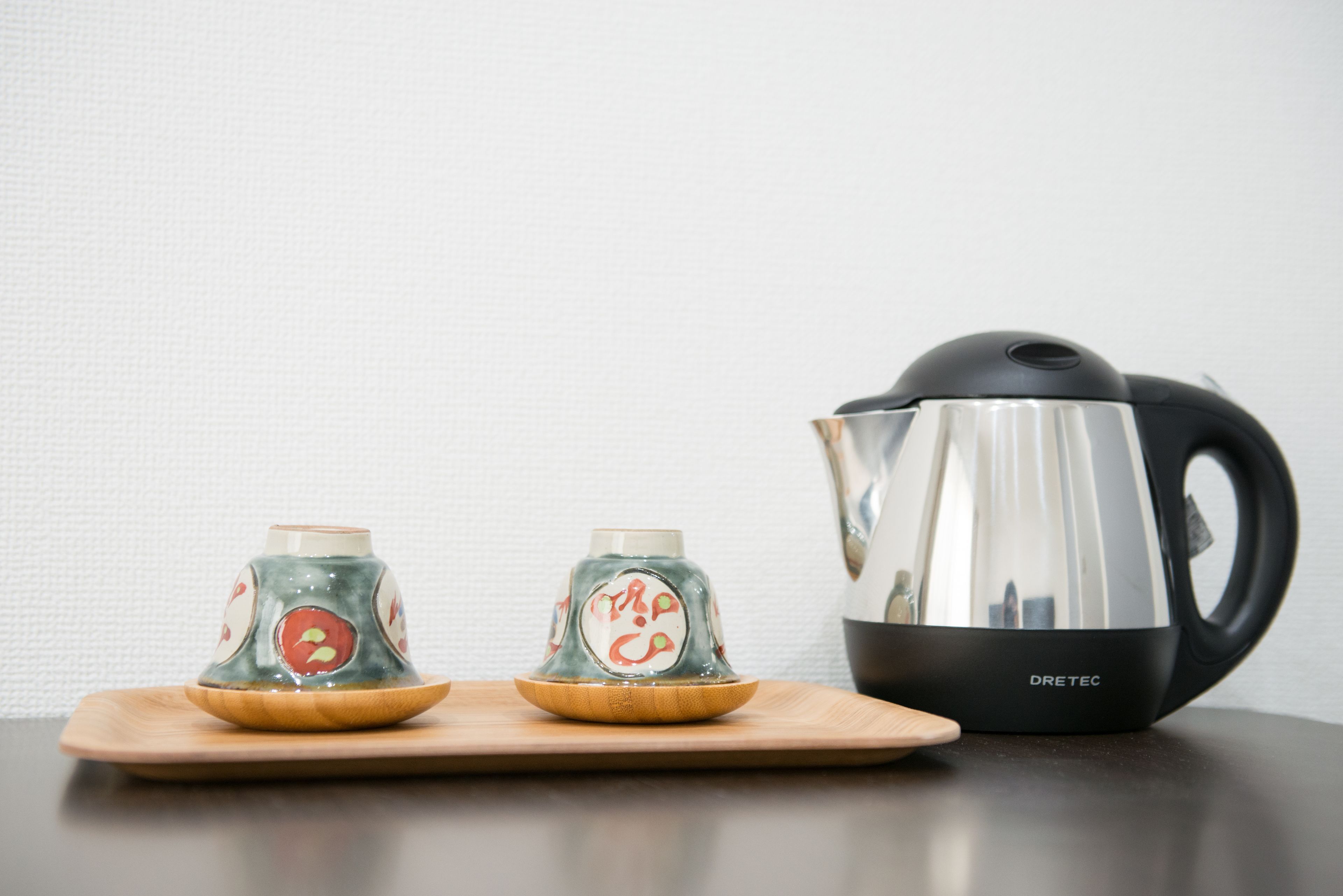 Two glass cups with lids on a wooden tray, a black electric kettle, and a wooden table.