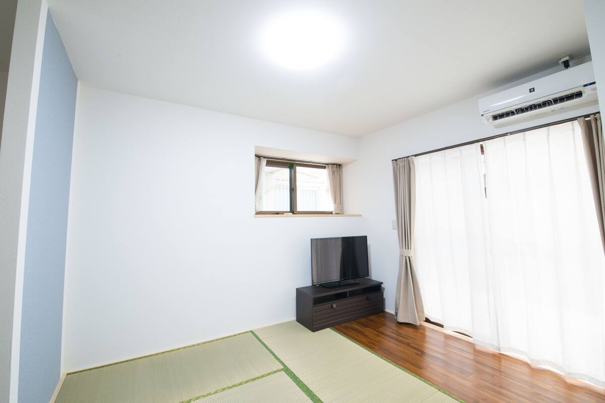 A room with a flat-screen TV on a dark wooden stand, a small window with curtains, and an air conditioning unit mounted on the wall.